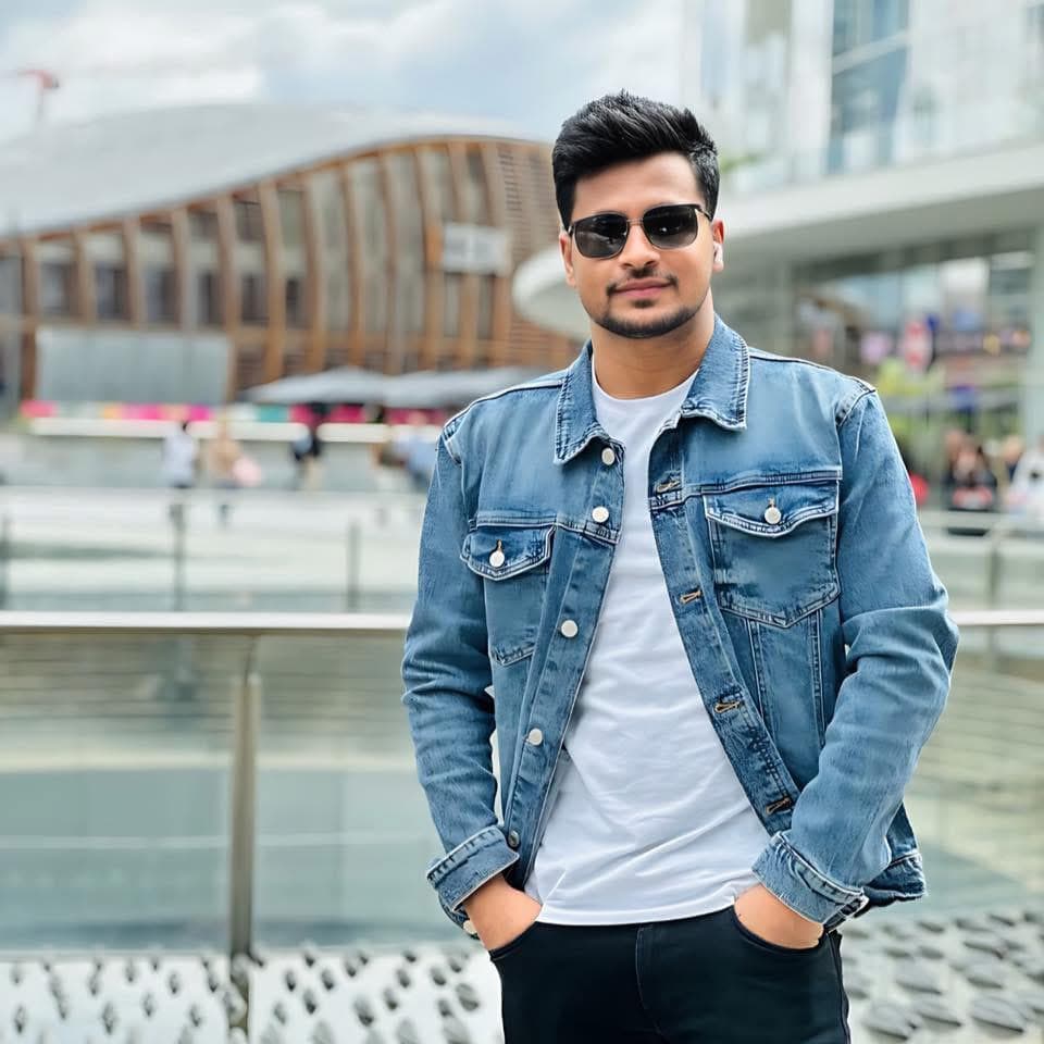 Man wearing a denim jacket and sunglasses posing outdoors against a blurred urban backdrop.